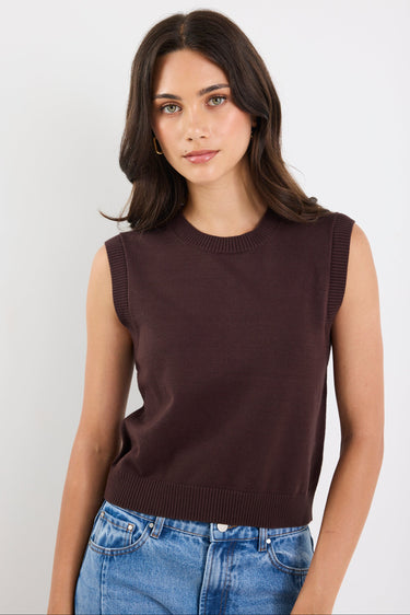 Woman wearing a brown sleeveless sweater and blue jeans against a white background