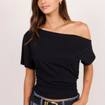 A woman modeling a black off-the-shoulder short-sleeve top paired with blue jeans and a black belt with a gold buckle, set against a plain light background.