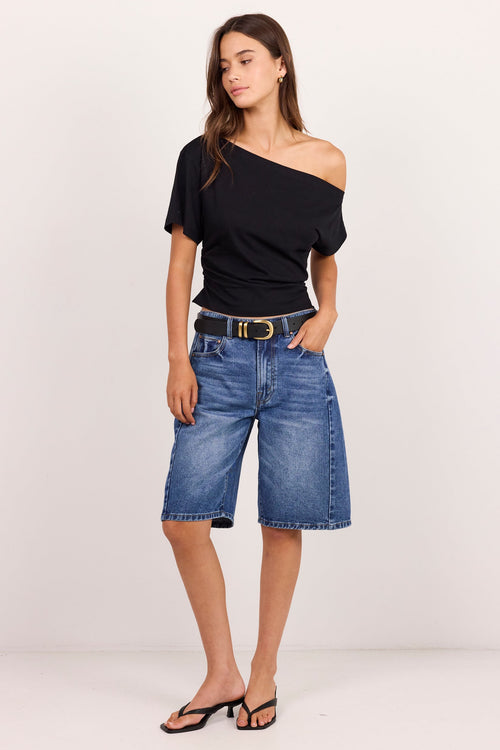A woman modeling a black off-the-shoulder short-sleeve top and blue denim cropped jeans with a black belt, paired with black heeled sandals, against a plain white studio background.