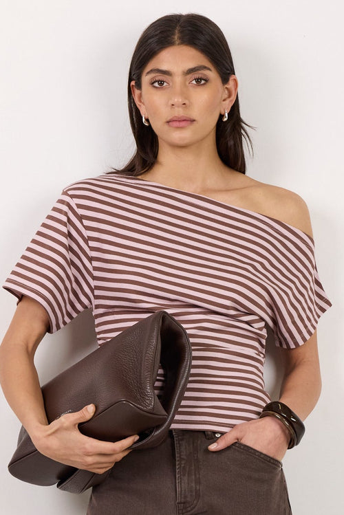 A woman stands against a plain off-white studio wall wearing a striped off-the-shoulder top, dark brown cropped trousers, and black heeled sandals, while holding a dark brown clutch.
