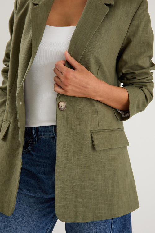 Person wearing an olive green blazer over a white top and blue jeans on a light gray background