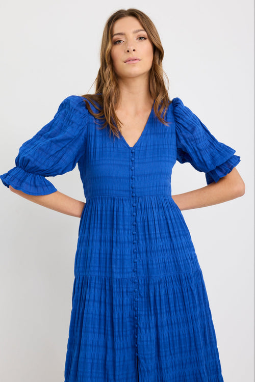 Woman wearing a bright azure blue tiered maxi dress with puff sleeves, button-front bodice, and v-neckline, styled with black woven flats.