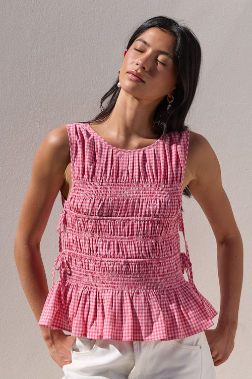 A woman wearing a pink and white gingham smocked sleeveless top with a ruffled hem, paired with white pants, posing against a neutral light gray wall.