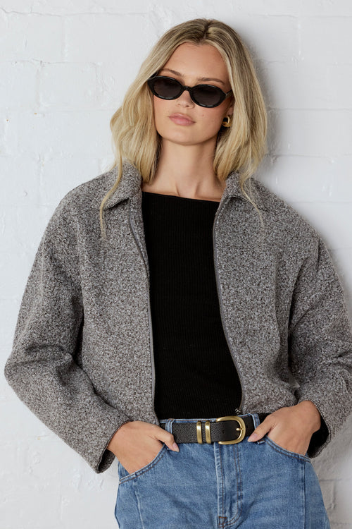 Woman wearing a gray jacket, black top, and blue jeans against a white wall.