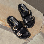 Black sandals with gold buckles on a stone surface