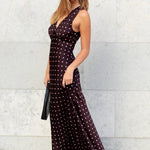 Woman in a dark dress with white polka dots standing against a light stone wall.