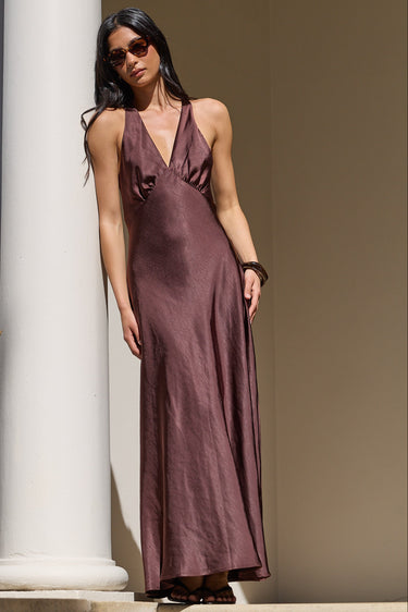 A woman wearing a sleeveless, floor-length mauve satin slip dress and sunglasses stands on stone steps beside white columns.