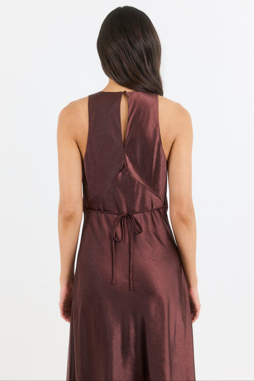 A woman viewed from the back wearing a sleeveless dark plum-brown dress with a tie waist and open back detail, against a plain light background.