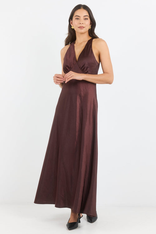 A woman modeling a sleeveless, floor-length wrap-style gown in a deep plum-brown satin fabric against a clean white studio background.