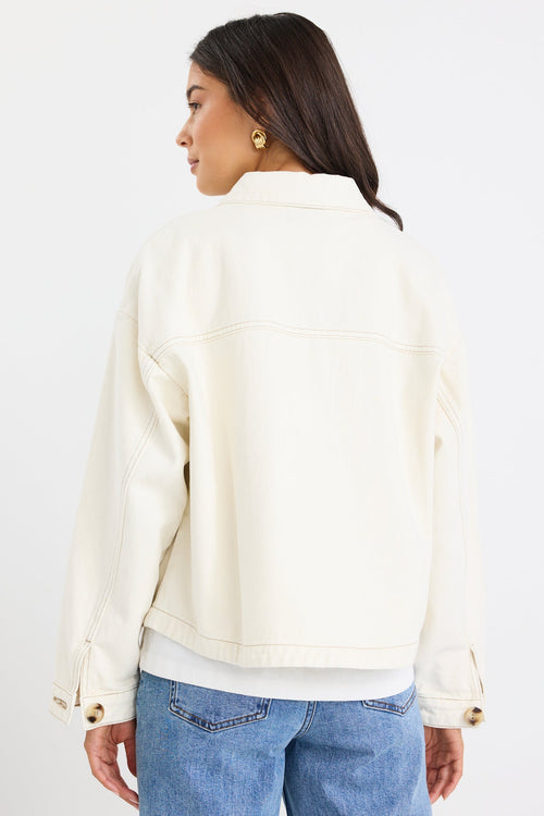 A woman shown from the back wearing a cream-colored denim jacket, a white t-shirt, and blue jeans against a plain white studio background.