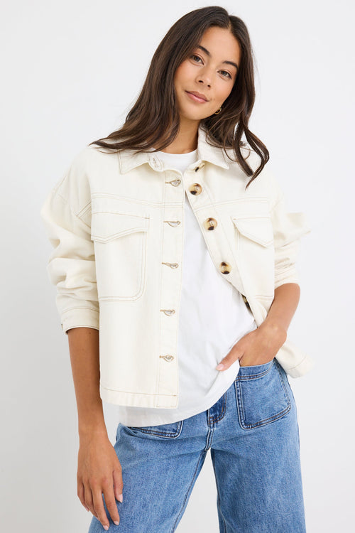 A woman modeling a cream-colored denim jacket with gold buttons over a white t-shirt and blue jeans, set against a plain white studio background.