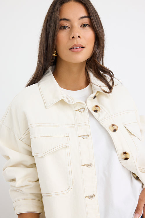 Woman wearing a relaxed fit off-white denim jacket with front flap pockets and tortoiseshell buttons, styled casually with jeans.