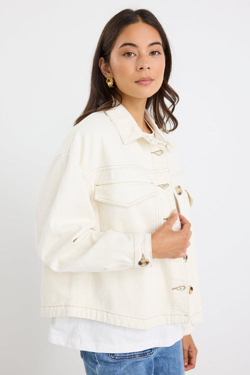 A woman wearing a cream-colored denim jacket with unique metal snap buttons, layered over a white t-shirt and blue jeans, standing against a plain white background.