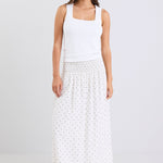 Woman wearing a white sleeveless top and long white skirt with black polka dots on a light gray background