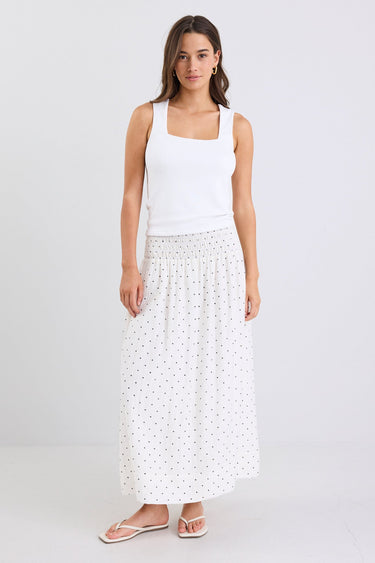Woman wearing a white sleeveless top and long white skirt with black polka dots on a light gray background