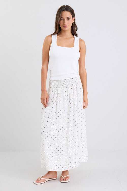 Woman wearing a white sleeveless top and long white skirt with black polka dots on a light gray background