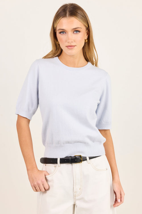 A woman modeling a light blue short-sleeve crewneck knit top, paired with cream-colored pants and a black belt, against a plain off-white studio background.