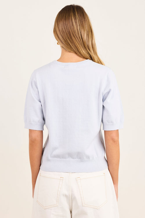 A woman viewed from behind wearing a light blue short-sleeve crewneck top and off-white pants, set against a plain cream background.