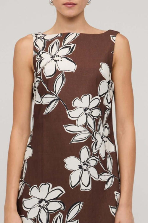 Brown sleeveless mini dress with a straight neckline and bold white floral print, styled with a loose A-line fit, worn by a model holding a woven handbag.