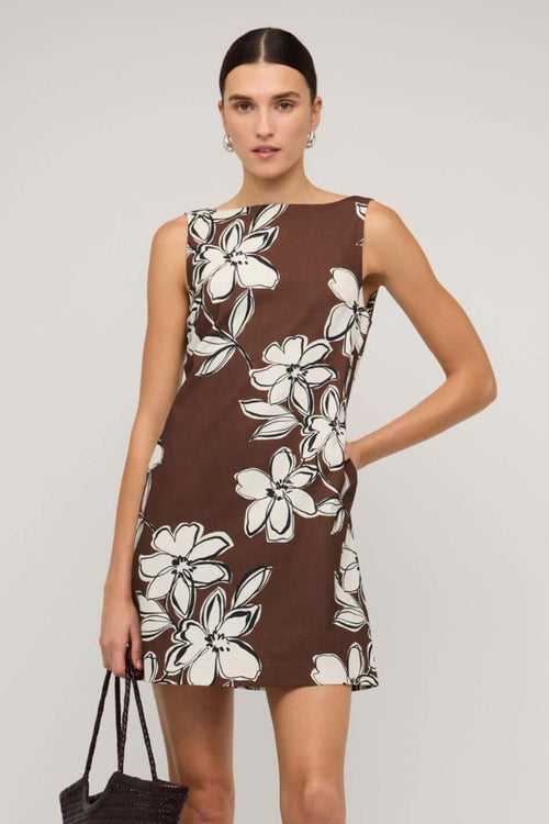 Brown sleeveless mini dress with a straight neckline and bold white floral print, styled with a loose A-line fit, worn by a model holding a woven handbag.