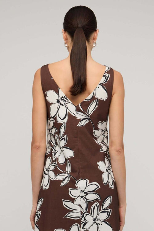 Brown sleeveless mini dress with a straight neckline and bold white floral print, styled with a loose A-line fit, worn by a model holding a woven handbag.