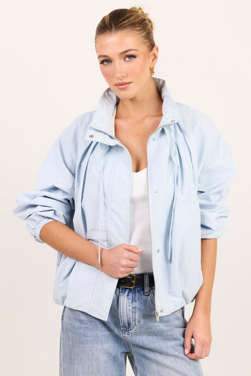 A woman modeling a light blue oversized button-up shirt jacket over a white tank top and light-wash jeans, photographed against a plain off-white studio background.
