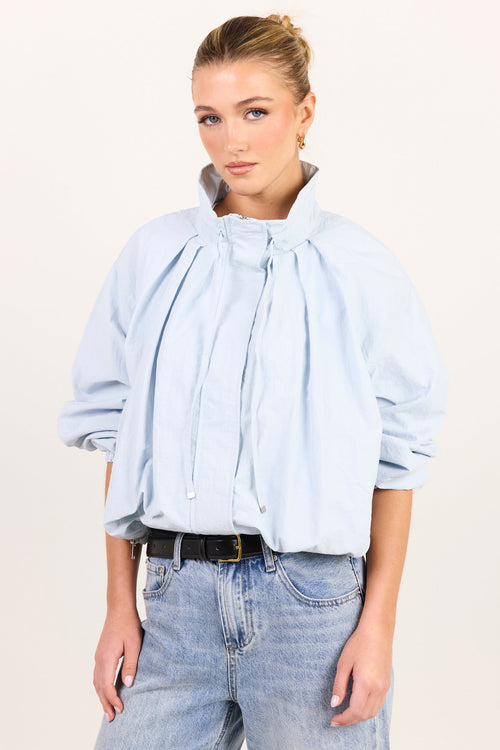 A woman modeling a light blue cropped blouse with dramatic gathered pleats and a high collar, paired with light-wash jeans and a black belt, against a plain off-white studio background.