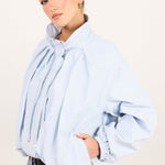 A woman wearing a light blue oversized long-sleeve blouse with a large gathered bow at the collar and elasticized cuffs, paired with light-wash blue jeans, against a plain off-white studio background.