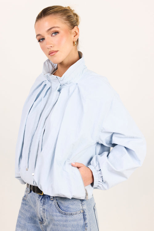 A woman wearing a light blue oversized long-sleeve blouse with a large gathered bow at the collar and elasticized cuffs, paired with light-wash blue jeans, against a plain off-white studio background.