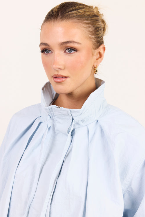 A close-up shot of a woman wearing a light blue high-neck blouse with dramatic gathered pleats and a high collar, set against a plain off-white background.