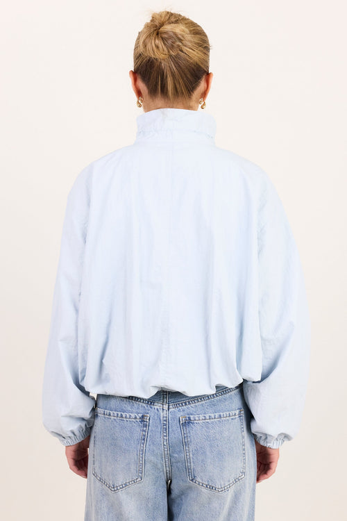 A person viewed from the back wearing a light blue high-neck long-sleeve shirt and light-wash blue jeans against a plain off-white studio background.