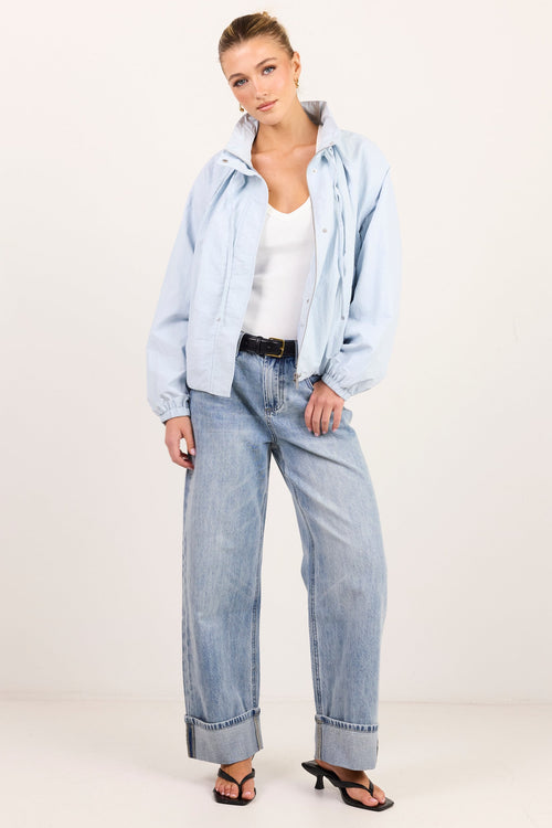 A woman modeling a light blue denim jacket and matching wide-leg jeans with a white tank top and black sandals, standing against a plain off-white studio background.