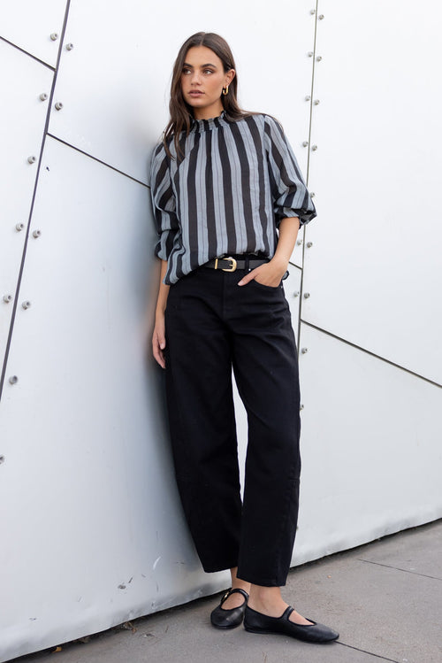 Woman in a striped shirt and black pants leaning against a modern architectural wall.