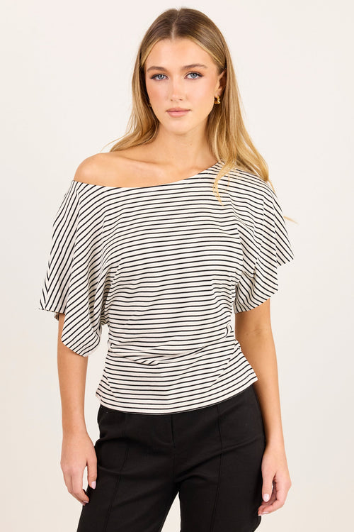 A woman modeling an off-the-shoulder top with black and white horizontal stripes, paired with black pants, against a plain light background.