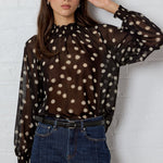 Woman wearing a sheer polka dot blouse and jeans against a white brick wall.