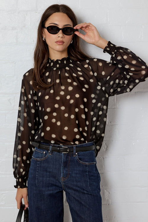 Woman wearing a sheer polka dot blouse and jeans against a white brick wall.