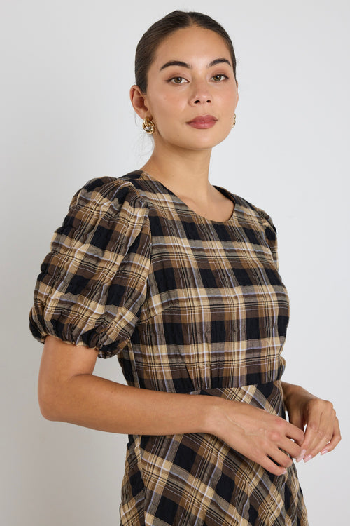 Woman wearing a caramel check maxi dress with balloon sleeves and flowing bias-cut skirt, styled with casual sneakers.