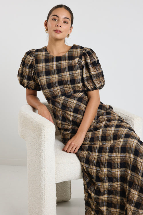 Woman wearing a caramel check maxi dress with balloon sleeves and flowing bias-cut skirt, styled with casual sneakers.