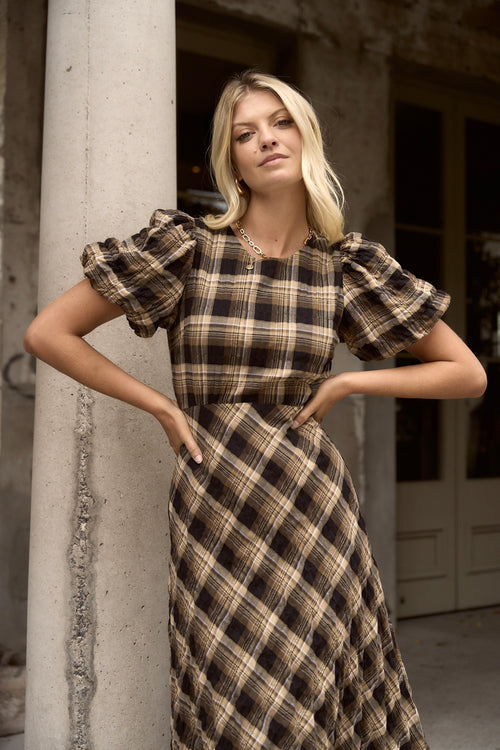 Woman wearing a caramel check maxi dress with balloon sleeves and flowing bias-cut skirt, styled with casual sneakers.