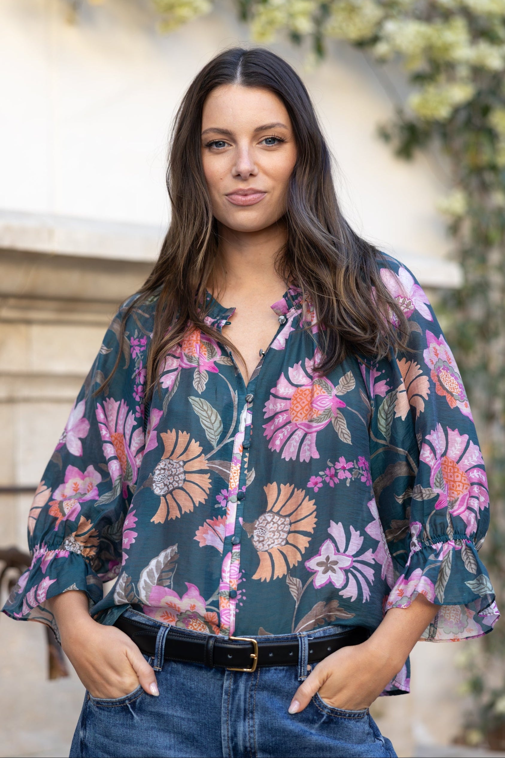 A woman wearing a dark teal floral blouse with pink, purple, and orange flowers, paired with blue jeans and a black belt, standing outdoors in front of a stone wall and greenery.