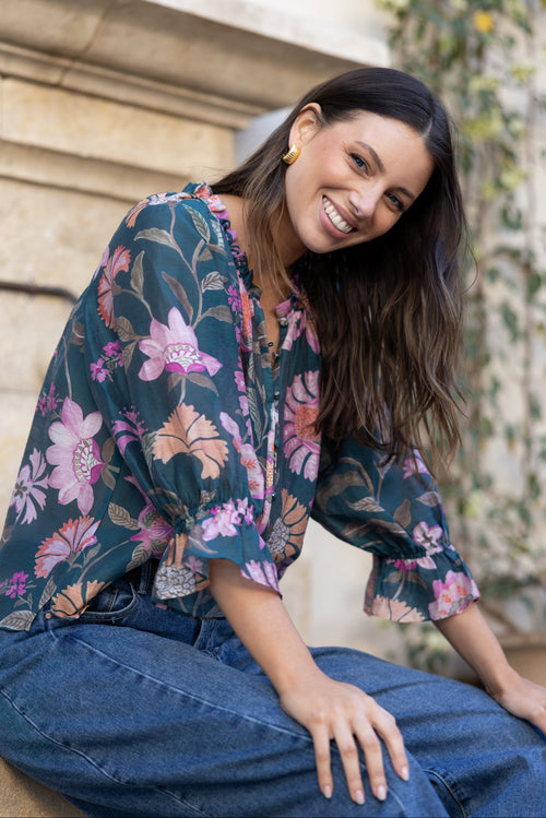 A woman wearing a dark teal floral blouse with pink and purple flowers, paired with wide-leg blue jeans, sitting outdoors on stone steps.