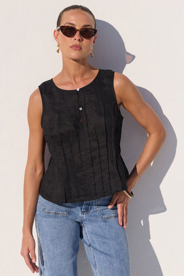 A woman wearing a black sleeveless top with vertical pleats and light blue jeans, standing against a white wall with a strong shadow.