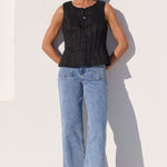 A woman stands against a white wall wearing a sleeveless black top, light blue wide-leg jeans, and black sandals.