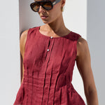A woman wearing a sleeveless, deep red linen peplum top with vertical pleats and a button detail, paired with beige trousers and sunglasses, standing against a white wall.