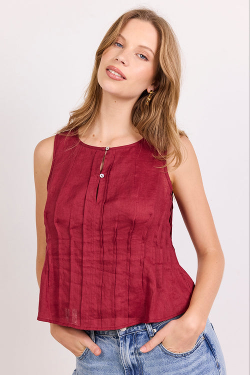 A woman wearing a sleeveless, deep burgundy top with a front zipper and button detail, paired with light-wash blue jeans, against a plain light gray background.