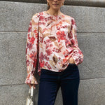  Woman wearing a blush floral top with flowy sleeves, paired with natural linen pants for an elegant and relaxed look.