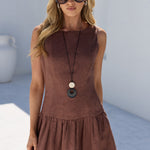 A woman modeling a sleeveless brown linen-look mini dress with a gathered waist and flared skirt, styled with a long pendant necklace and sunglasses in a bright white-walled outdoor setting.