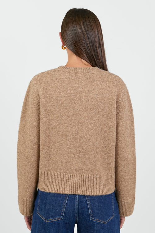 A woman wearing a soft, biscuit-colored crew neck jumper with long sleeves and ribbed cuffs, paired with dark blue high-waisted jeans.