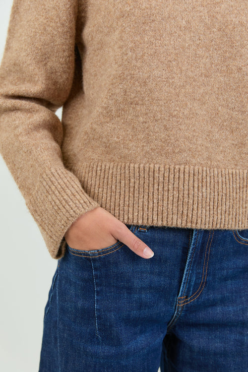 A woman wearing a soft, biscuit-colored crew neck jumper with long sleeves and ribbed cuffs, paired with dark blue high-waisted jeans.