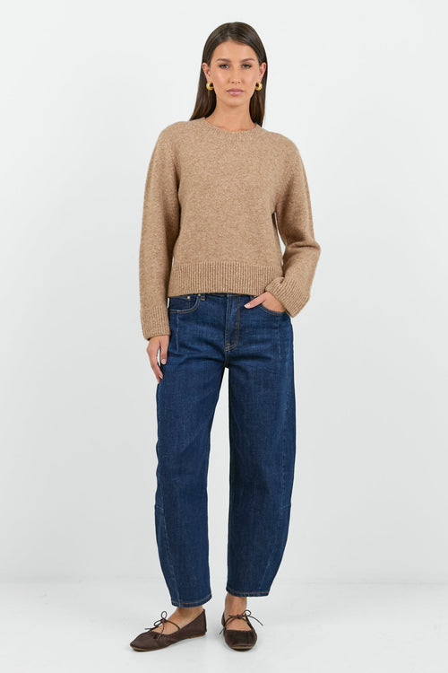 A woman wearing a soft, biscuit-colored crew neck jumper with long sleeves and ribbed cuffs, paired with dark blue high-waisted jeans.